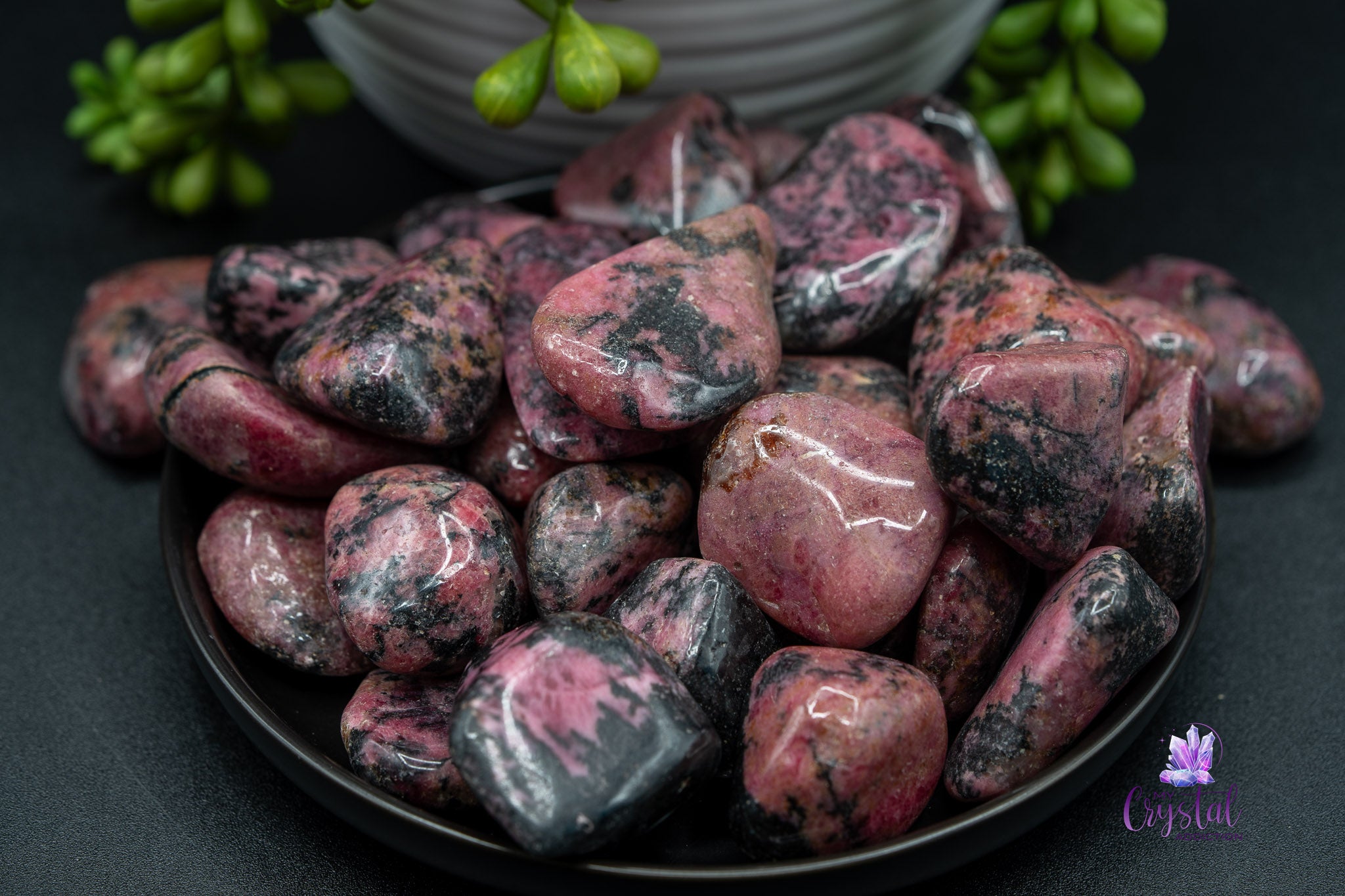 Polished rhodonite tumbled stones in a black bowl, succulent plant in background
