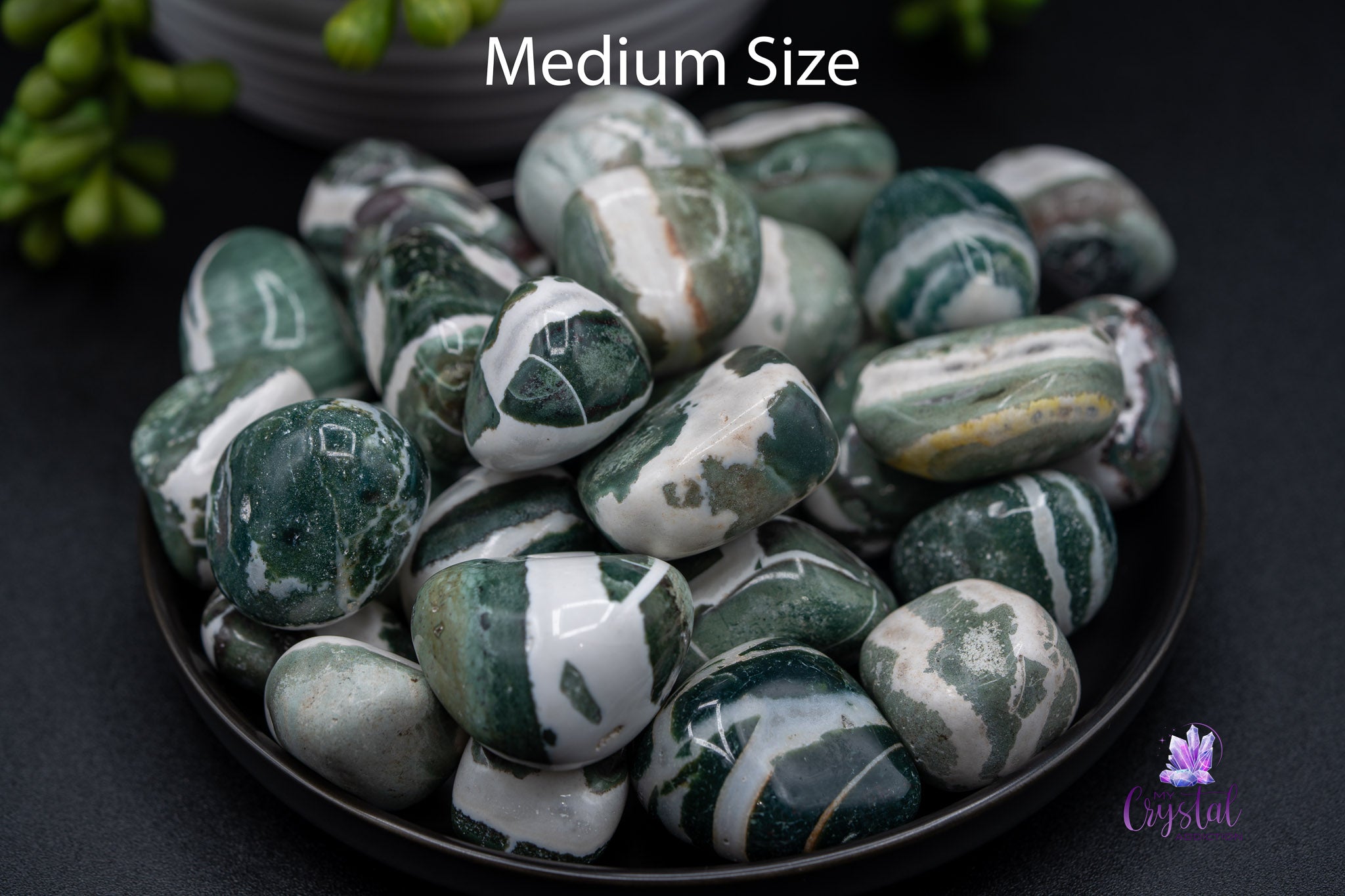 Medium size tumbled green and white moss agate stones in a black bowl on dark surface