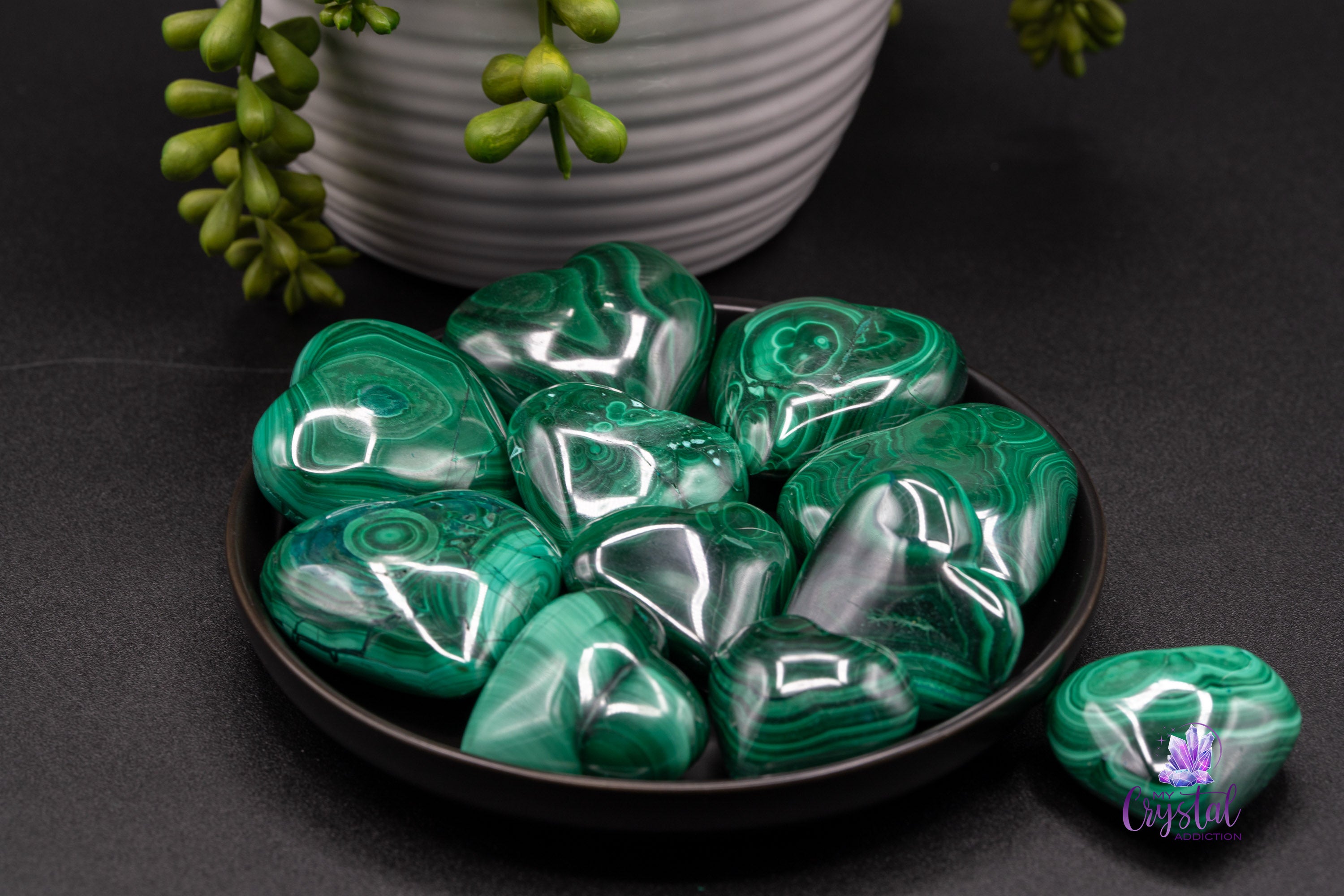 Polished green malachite heart carvings in a black dish, showing glossy surfaces and layered green bands.